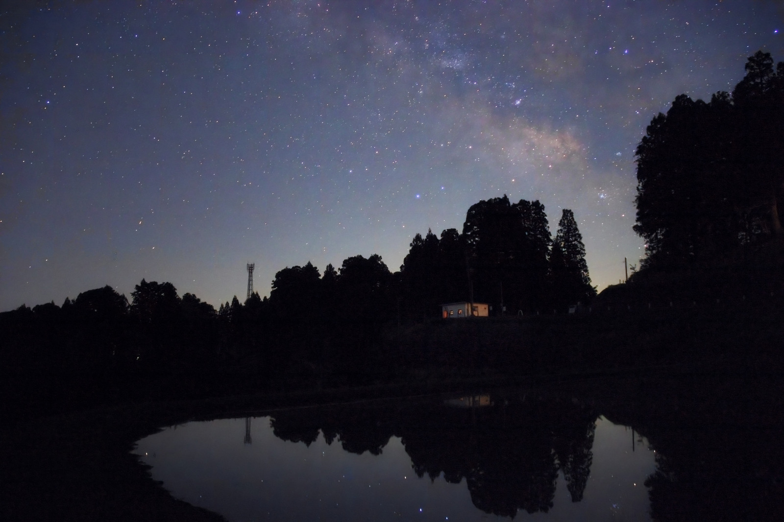 Edited photograph: A diagonal band of the Milky Way above silhouetted trees and a warmly lit house, reflected in a woodland pond.