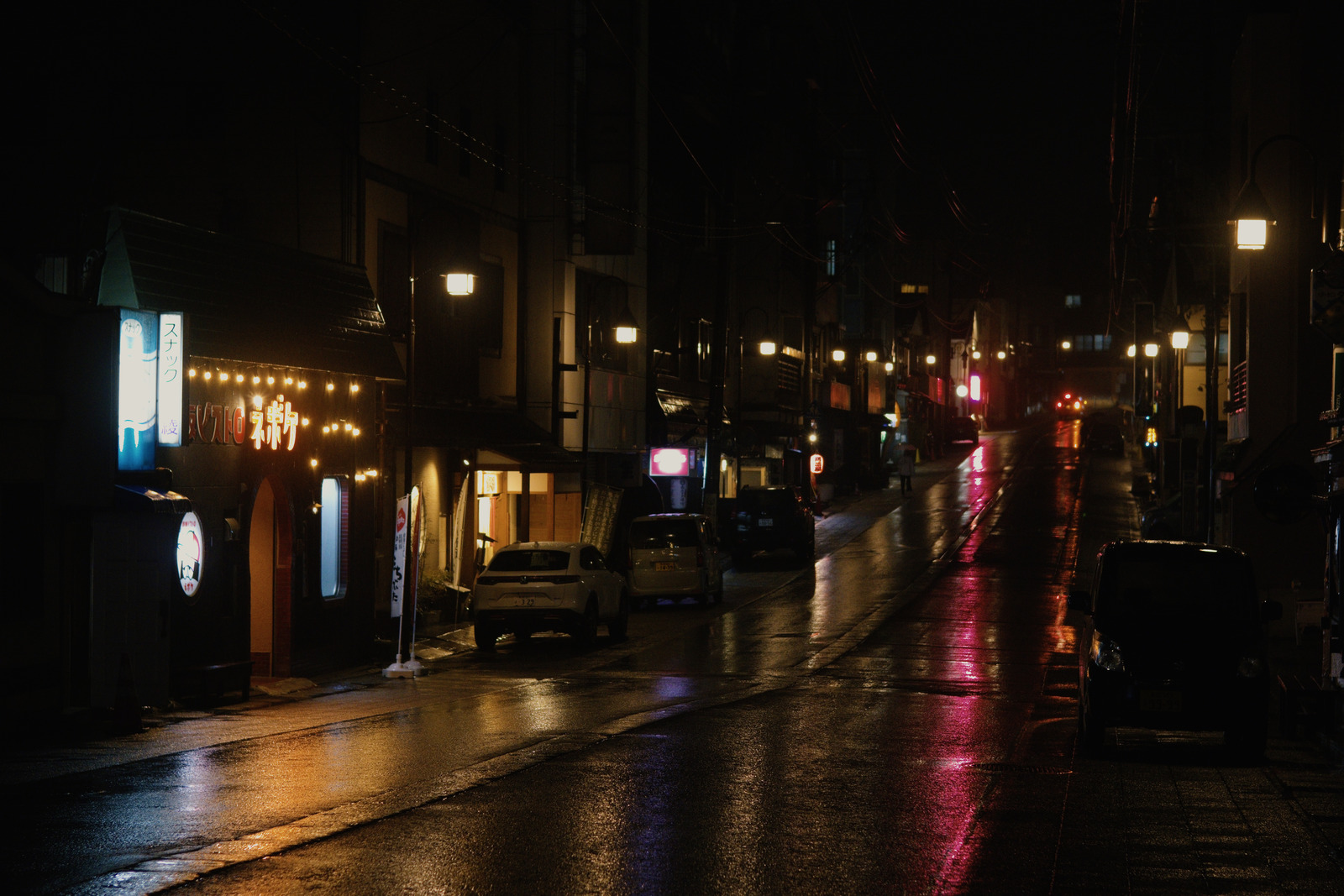 After Hours preset applied to the same base rendering: Warm streetlights and coloured shop signs reflected along a dark wet street with parked cars.