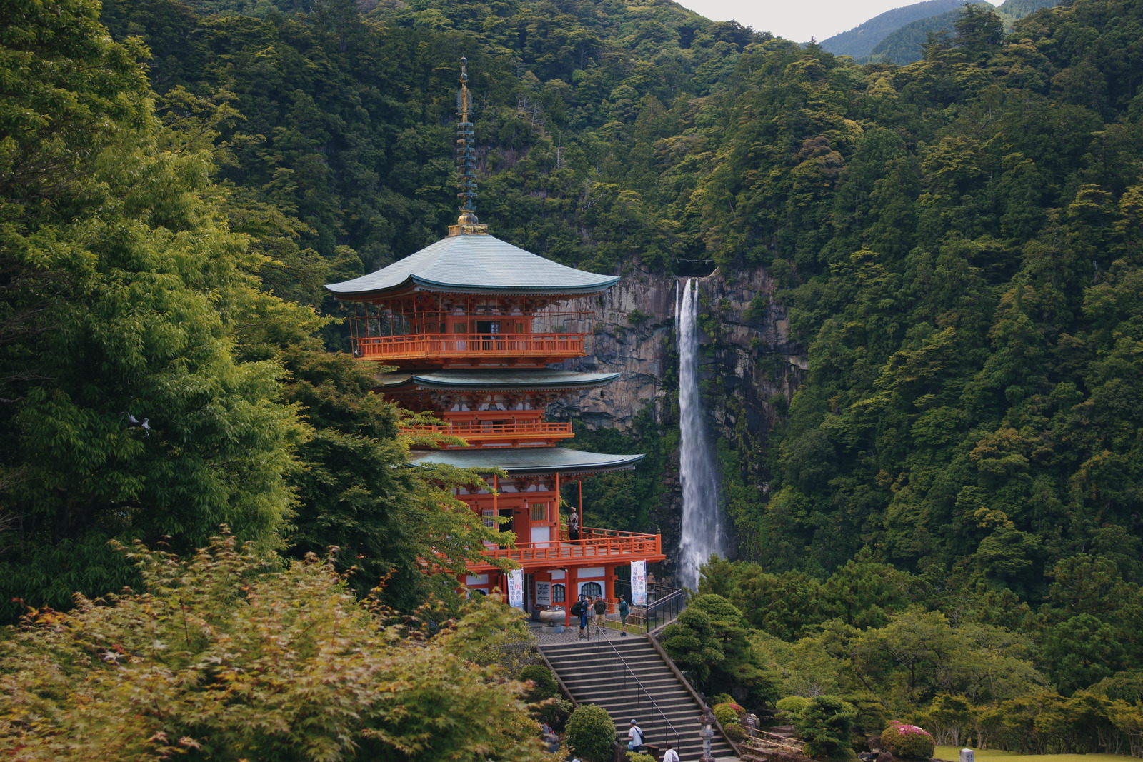 A vermilion pagoda beside a narrow waterfall, surrounded by a densely wooded hillside.